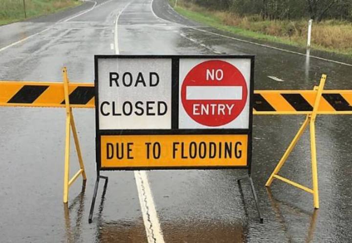 Yellow sign that says 'water on road' sits in middle of a flooded rural road flanked by black and yellow-striped barricades